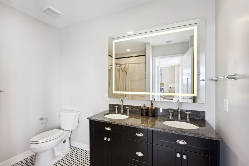 Modern bathroom with a double-sink dark vanity, granite countertop, large illuminated mirror, white toilet, towel bar on the wall, and a shower with a curtain. White walls and black-and-white patterned floor tiles.