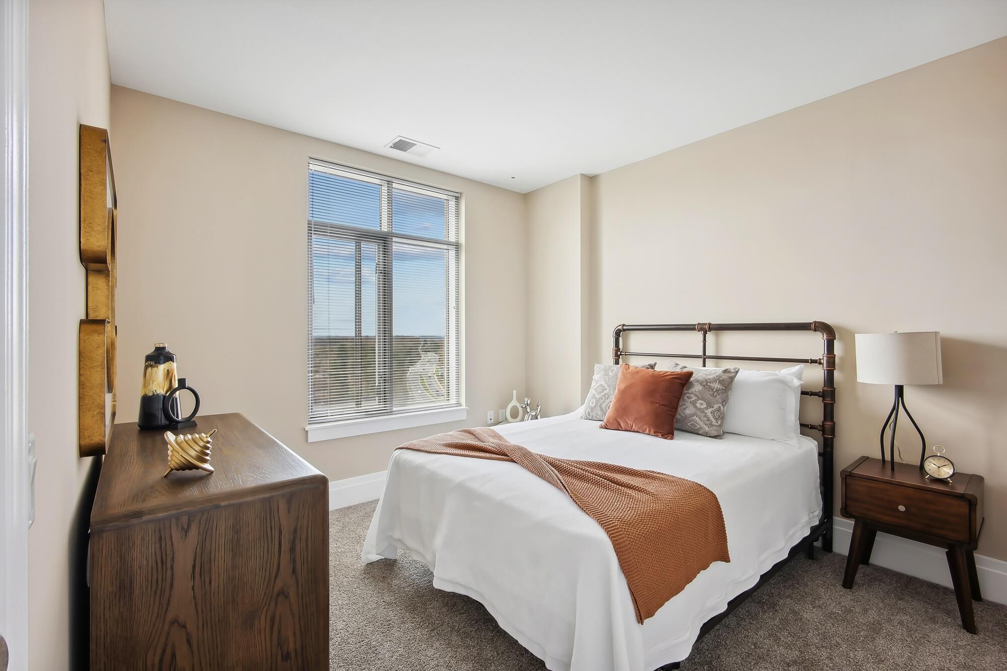 A cozy bedroom with a metal-frame bed, white bedding, and brown throw pillows. There is a wooden nightstand with a lamp and clock, a dresser with decor items, and a large window letting in natural light.