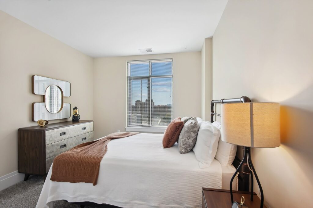 A modern bedroom with a bed covered in white linens and brown accents, a brown dresser with a decorative mirror above it, a bedside table with a lamp, and a window letting in natural light.