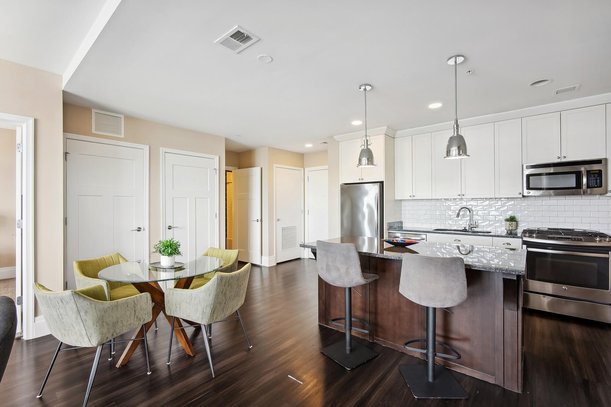 Modern kitchen with white cabinets, stainless steel appliances, a dark wood island with two gray barstools, and a round glass dining table with four green chairs on dark wood floors.