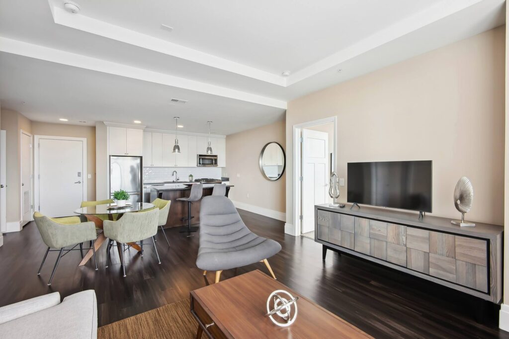 Modern open-concept living room and kitchen with dark wood floors, light walls, a TV on a wooden stand, round dining table with green chairs, and stainless steel kitchen appliances.