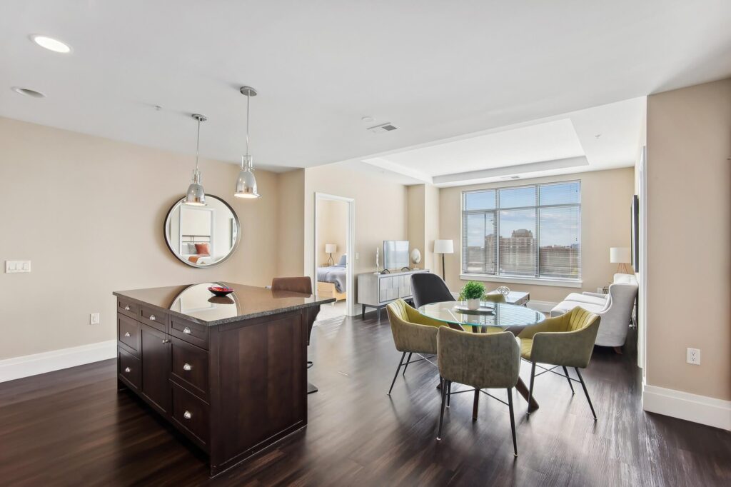 Modern apartment interior with an open kitchen island, round dining table and chairs, living area with a sofa and TV, large window with city view, and a mirror on the wall reflecting another room.