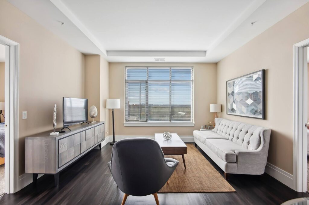 Modern living room with beige walls, a large window, white sofa, black accent chair, wooden coffee table, TV on a sleek stand, and floor lamps; natural light fills the space.