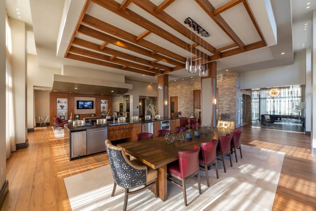 A spacious, modern open-concept kitchen and dining area with wood floors, a large dining table with red chairs, a kitchen island, built-in shelves, and a coffered wooden ceiling. Natural light fills the room.