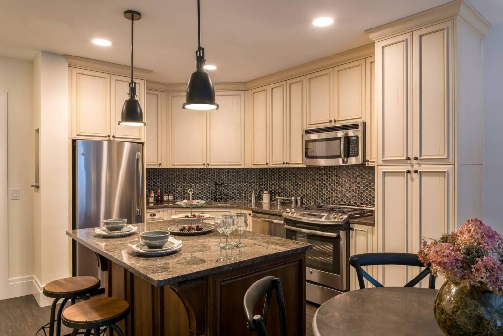 Modern kitchen with cream cabinets, stainless steel appliances, mosaic tile backsplash, and a granite island set for four. Pendant lights hang above the island, and a round dining table with flowers is in the foreground.