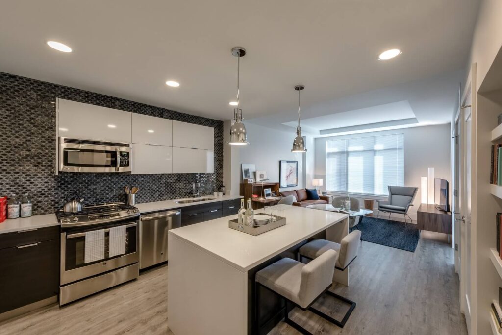 Modern kitchen and living area with stainless steel appliances, white cabinets, a large island with bar stools, pendant lighting, and a cozy sitting area with a sofa, armchair, and large windows letting in natural light.