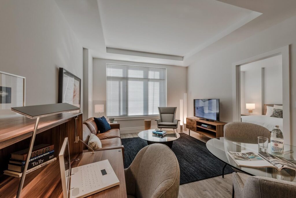 Modern living space with a desk, laptop, sofa, armchair, coffee table, TV, and a separate bedroom. Large window lets in natural light, and neutral décor with wood accents creates a cozy, minimalist atmosphere.