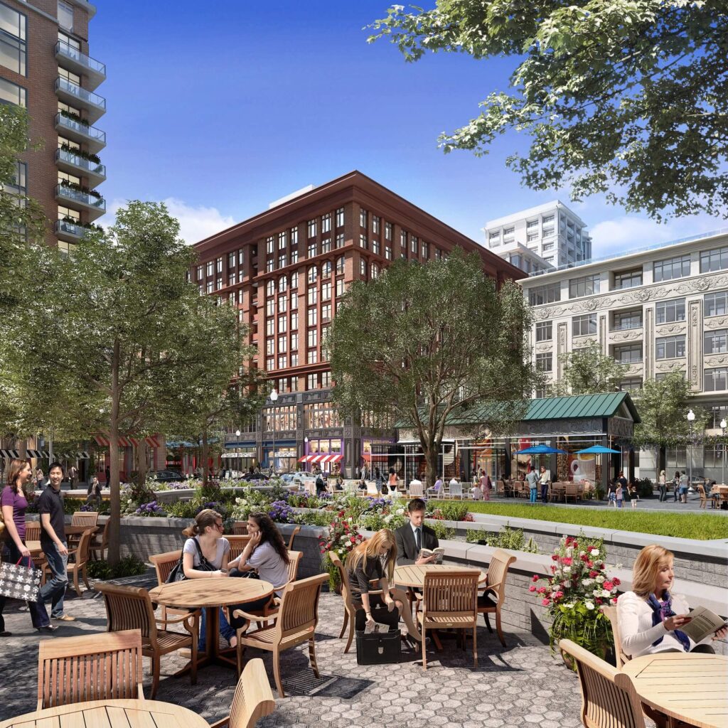 People sit at outdoor café tables in a lively urban plaza surrounded by trees, flowers, and tall buildings under a bright blue sky. Some people are eating, talking, or walking along the wide sidewalk.