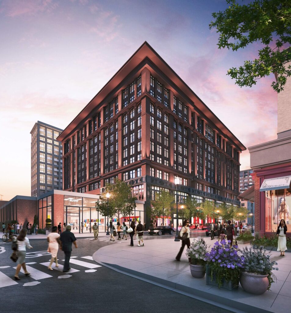 A modern red-brick building with large windows and shops at street level is surrounded by people walking on a busy urban sidewalk at sunset. Trees and flowers decorate the vibrant city scene.