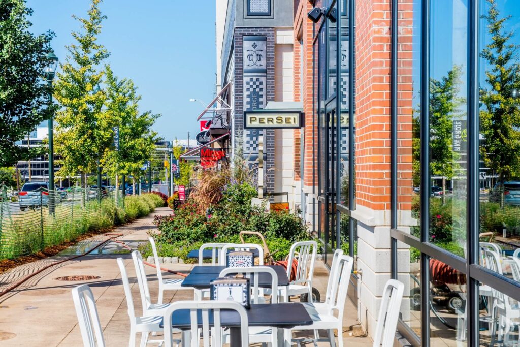 Outdoor cafe seating with white chairs and black tables on a sunny sidewalk next to a brick building with a PER SEI sign. Green trees and a street are visible in the background.