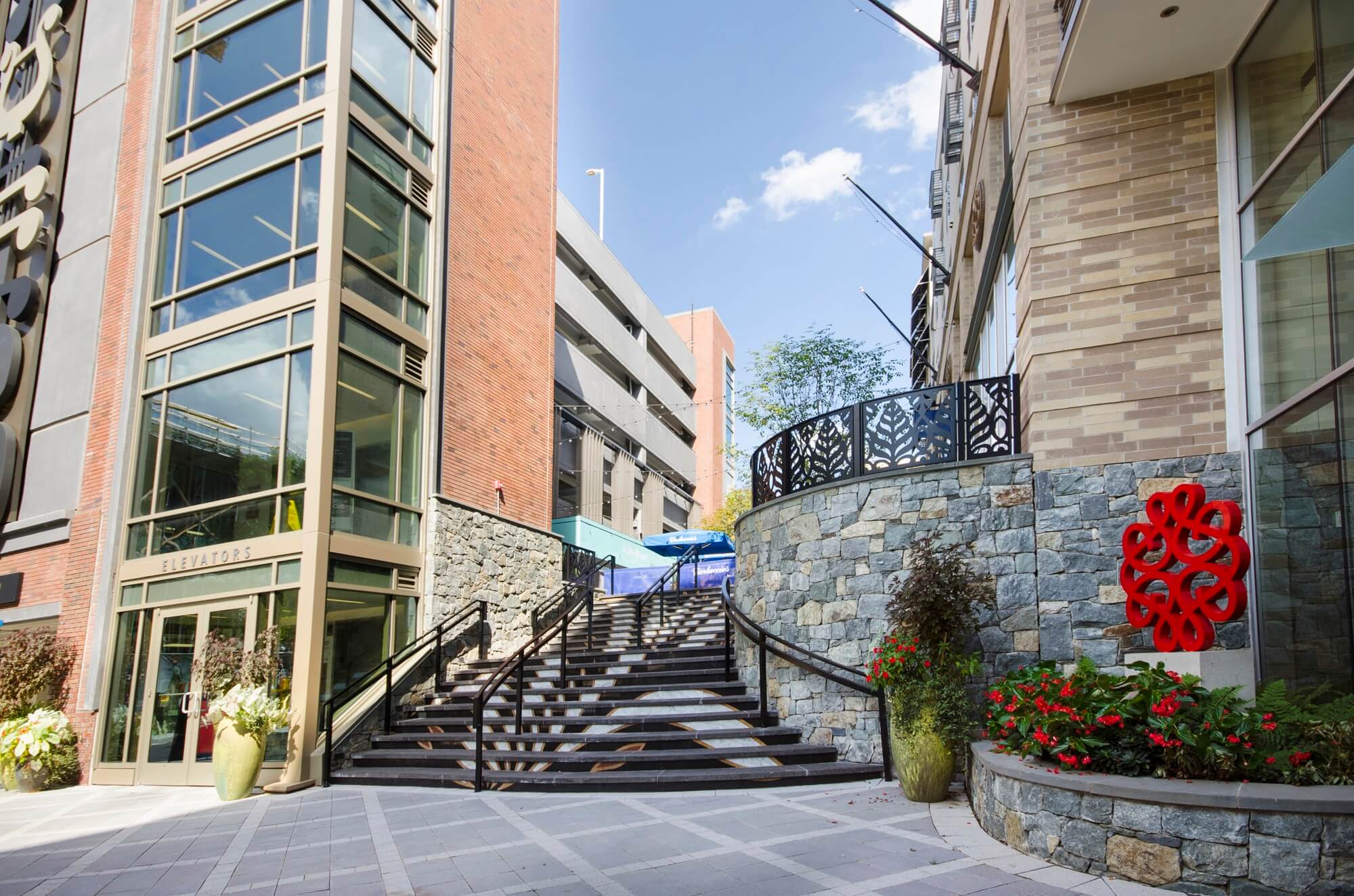 Outdoor steps lead between two modern buildings with glass, brick, and stone facades. Potted plants and a bright red abstract sculpture are visible near the entrance. Blue sky and greenery are seen in the background.