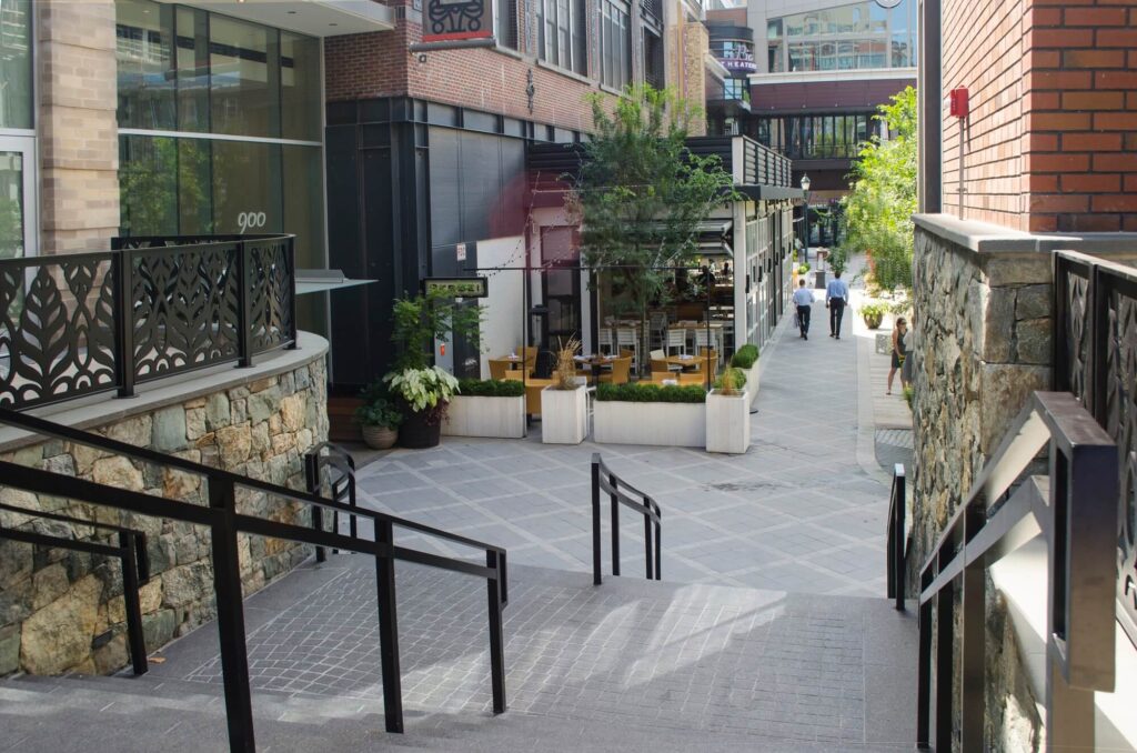 A modern outdoor urban walkway with stone walls, black railings, potted plants, and a few people walking in the distance near restaurant seating and storefronts.