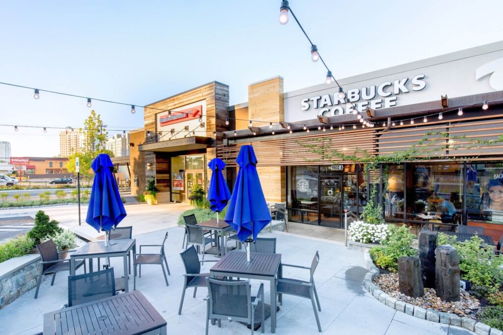 Outdoor seating area with tables, chairs, and closed blue umbrellas in front of a Starbucks Coffee shop, featuring string lights above and a modern, urban setting in the background.