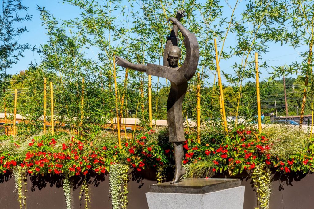 A bronze statue of a person with outstretched arms stands on a pedestal, surrounded by green bamboo and red flowers on a sunny day.