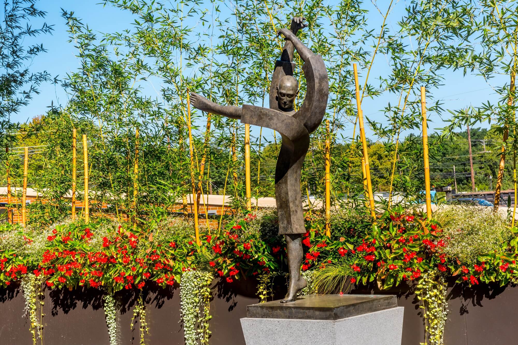 A bronze statue of a person with outstretched arms stands on a pedestal, surrounded by green bamboo and red flowers on a sunny day.