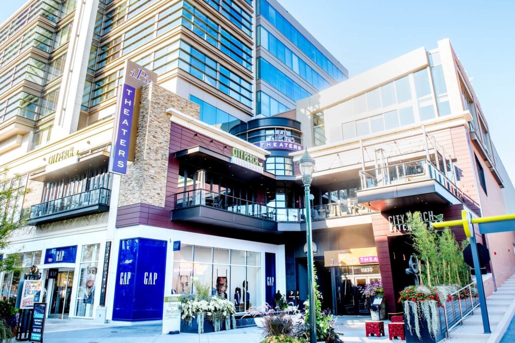 A modern outdoor shopping center with stores like GAP and a movie theater, featuring large glass windows, signage, and people walking by on a sunny day.