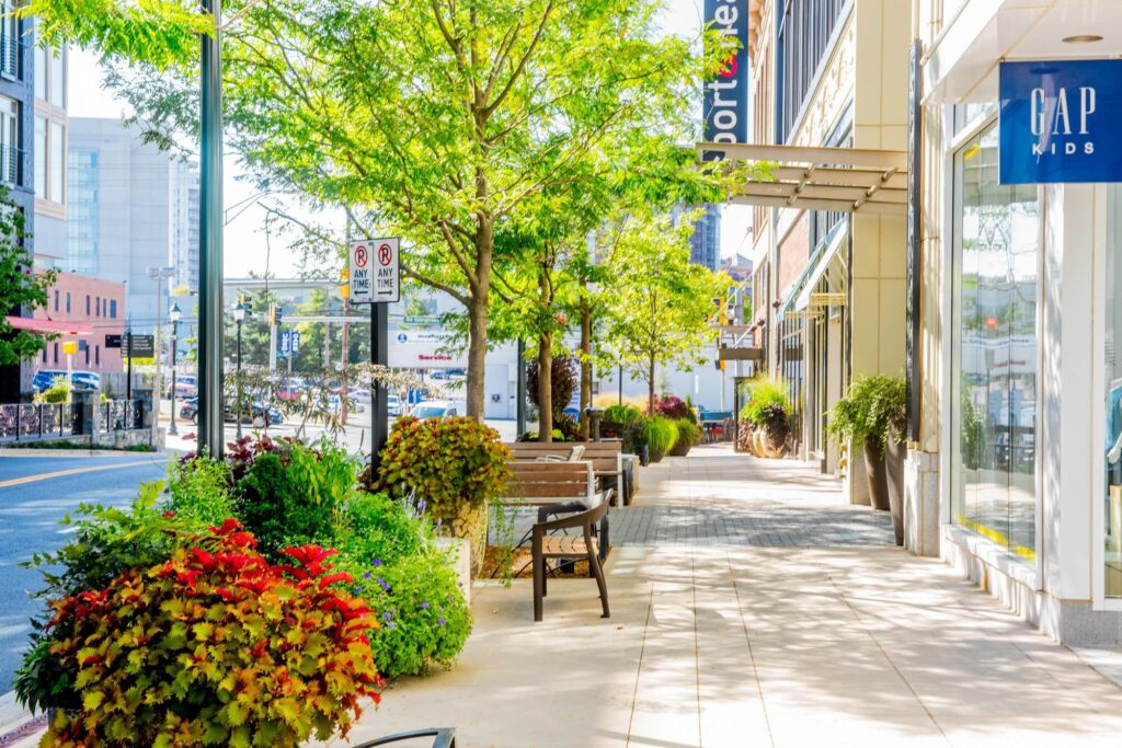 A sunlit city sidewalk lined with green trees, potted plants, benches, and storefronts, including a GAP Kids store. The scene is bright, clean, and inviting, with light traffic and modern buildings in the background.