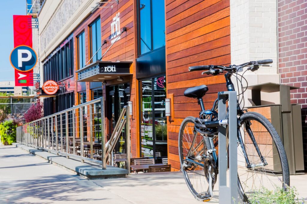 A black bicycle is locked to a bike rack on a sunny sidewalk outside a modern restaurant with wood paneling. A parking sign and outdoor seating are visible, along with a metal railing and storefront windows.