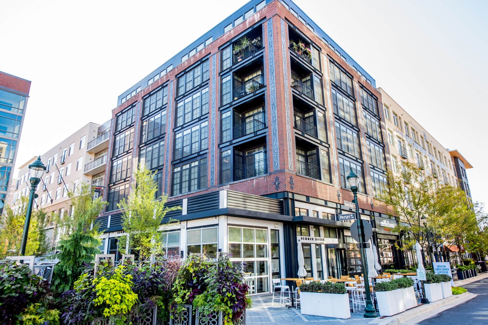 A modern, multi-story brick building with large windows and balconies sits on a city street corner, featuring a ground-floor café with outdoor seating and surrounded by trees and greenery.