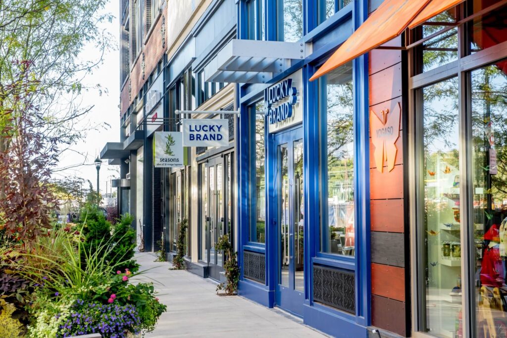 A row of modern retail storefronts, including Lucky Brand, with large windows, blue trim, and greenery along the sidewalk on a sunny day.