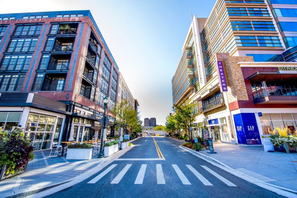 A wide street runs between modern multi-story buildings with shops, restaurants, and a theatre. Trees and planters line the sidewalks under a clear blue sky, giving a vibrant, urban feel.