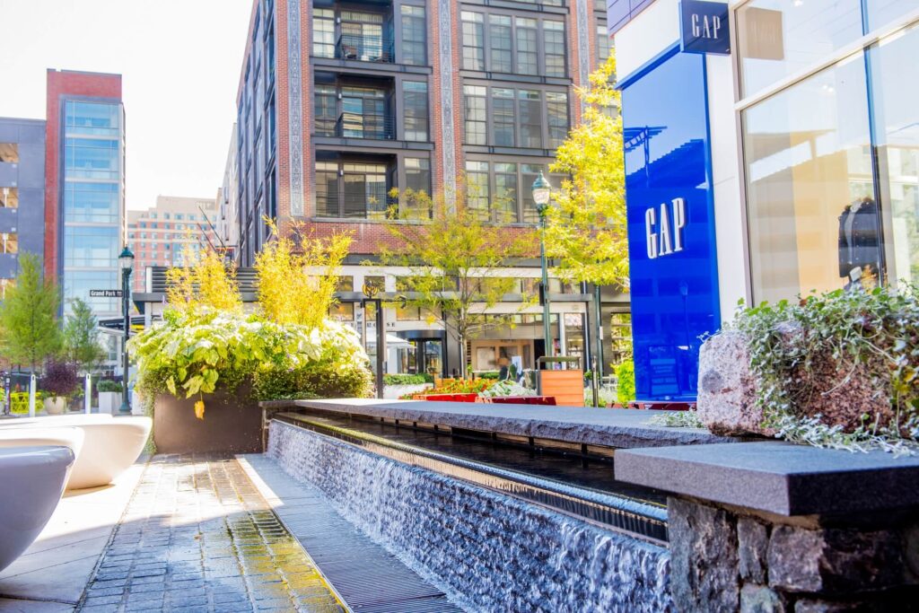 A modern urban plaza featuring a low stone fountain with cascading water, lush planters, benches, and surrounding buildings, including a brightly colored GAP storefront on the right.