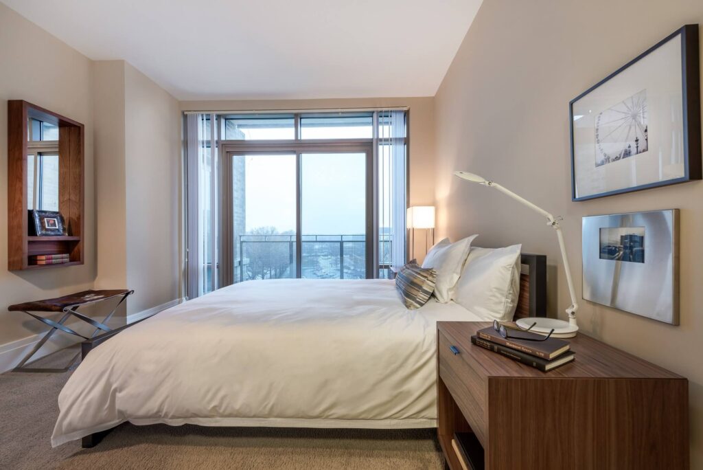 A modern bedroom with a large bed, white bedding, a wooden nightstand with books and a lamp, a wall mirror, and floor-to-ceiling windows letting in natural light with a view of trees outside.