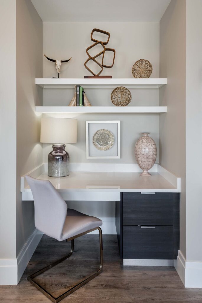 A modern, small home office nook with a white built-in desk, dark drawers, a beige chair, two shelves with decorative items, a lamp, books, and wall art. The space has neutral tones and a minimalist design.