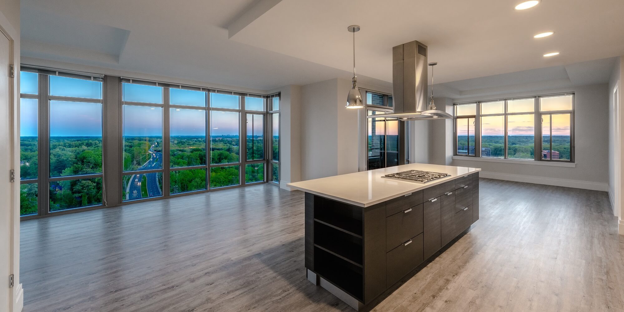 Modern open-concept apartment with large floor-to-ceiling windows, a central island kitchen, light wood floors, and expansive views of greenery and a sunset sky.