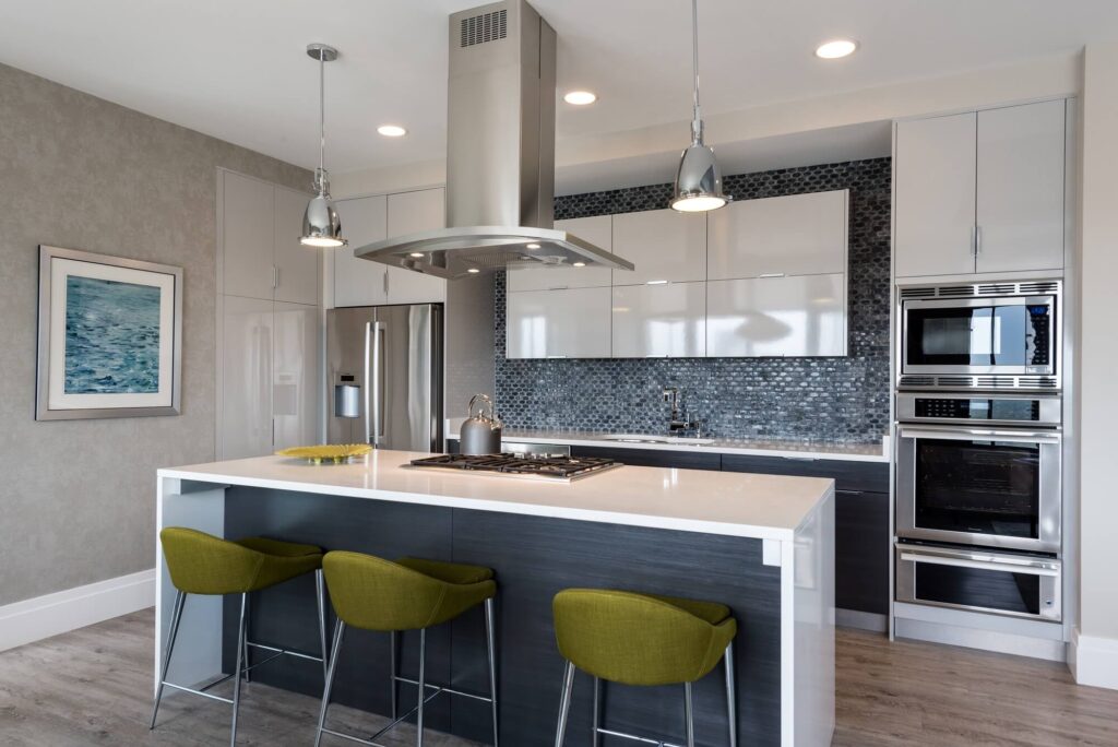 Modern kitchen with a white island, three green barstools, stainless steel appliances, mosaic tile backsplash, pendant lights, and a kettle on the stove. A framed picture hangs on the wall to the left.