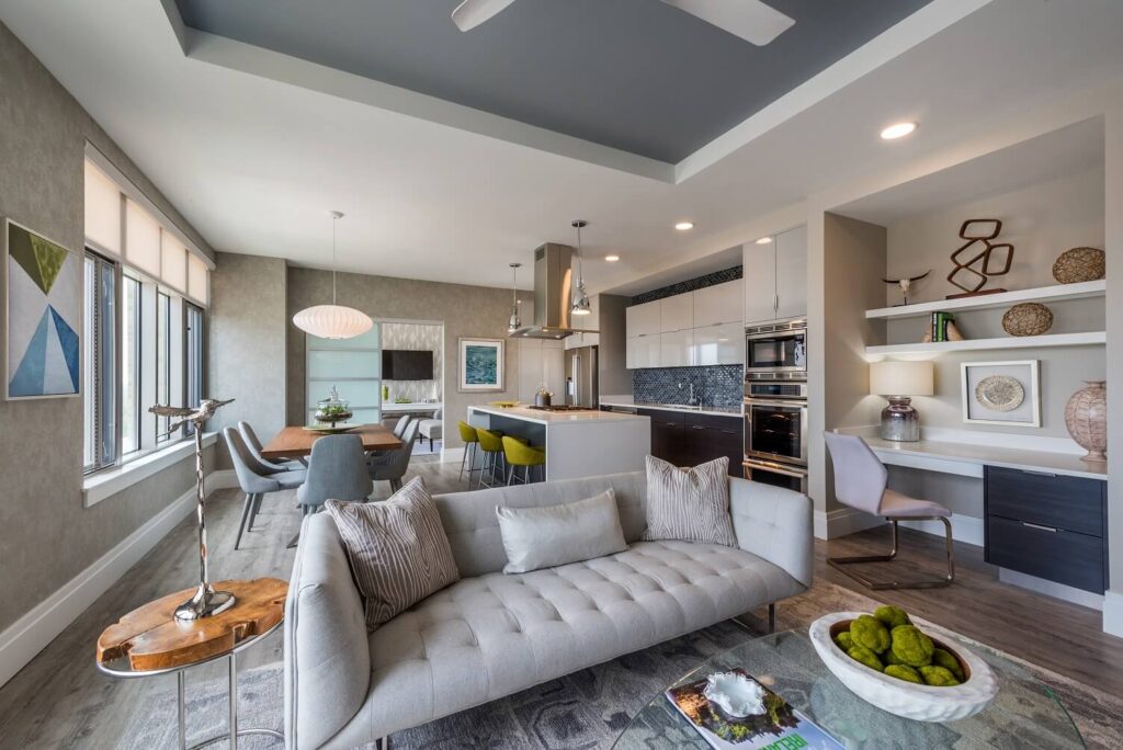 Modern open-concept living and dining area with a gray sofa, glass dining table, green chairs, built-in shelves, and contemporary decor. Large windows let in natural light, and the kitchen is in the background.