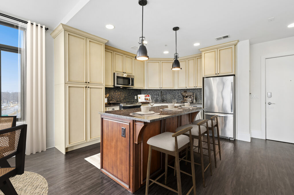 Modern kitchen with cream-colored cabinets, stainless steel appliances, a dark wood island with granite countertop, three bar stools, pendant lighting, and a window with a city view.