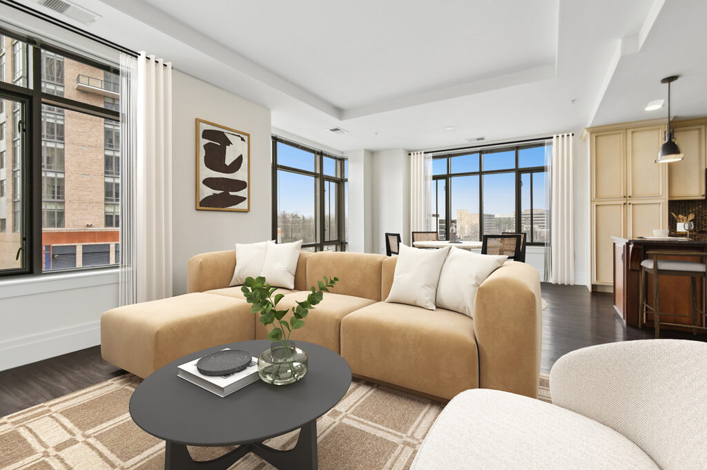 A modern living room with large windows, beige sofas, a black round coffee table with plants, abstract wall art, light curtains, and a view of city buildings outside. The kitchen and dining area are visible in the background.