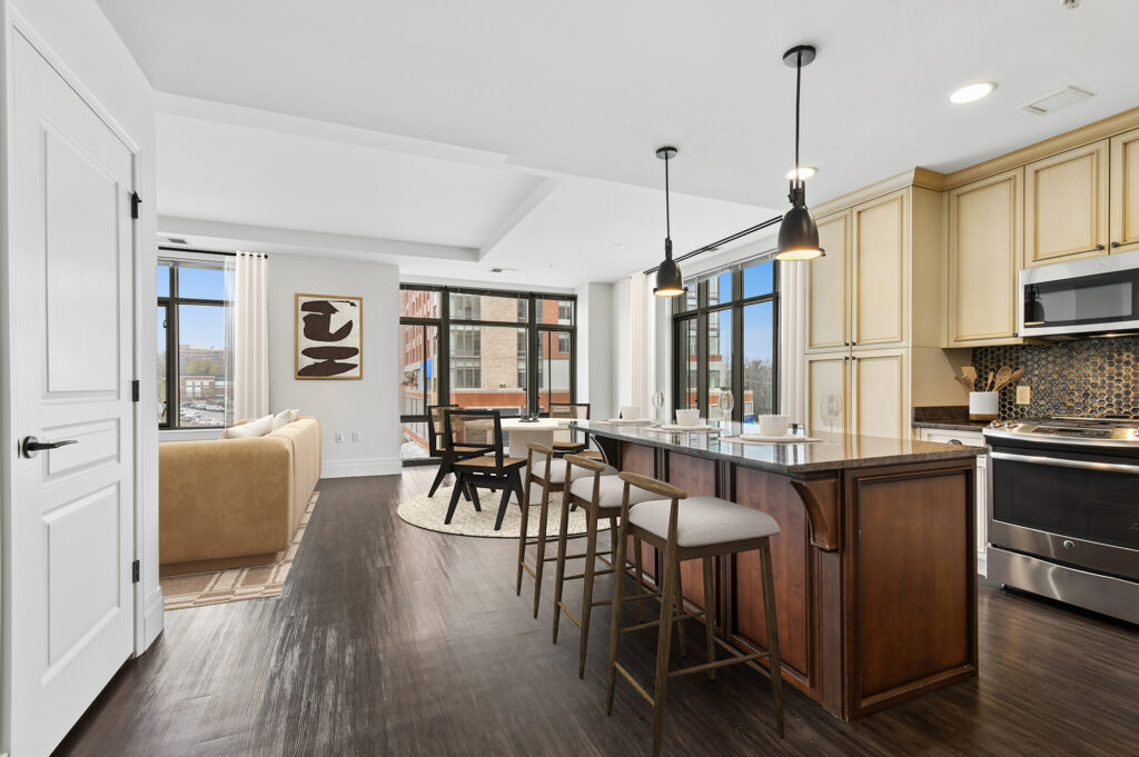 Modern open-concept kitchen and living area with large windows, neutral furnishings, an island with barstools, pendant lighting, beige cabinets, and a stainless steel oven. Artwork and a beige sofa are visible in the background.