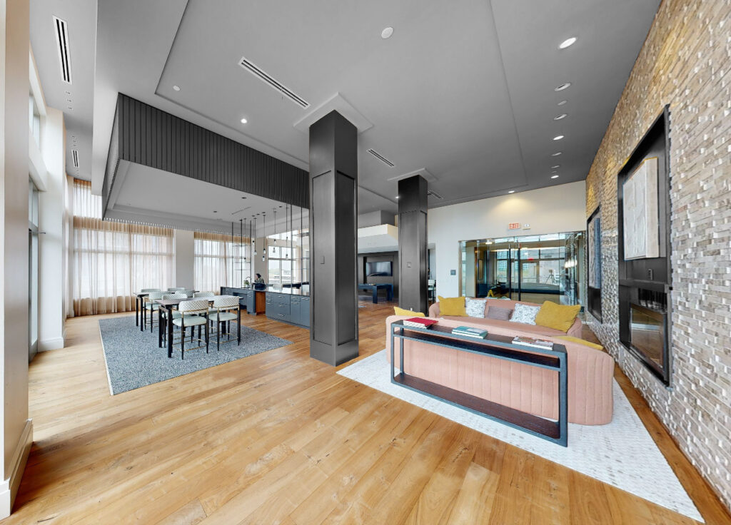 Modern, open-concept lounge with wood flooring, a dining area with a long table and chairs, a cozy seating area with a sofa and colorful pillows, large windows, and a stone accent wall with mounted TVs.