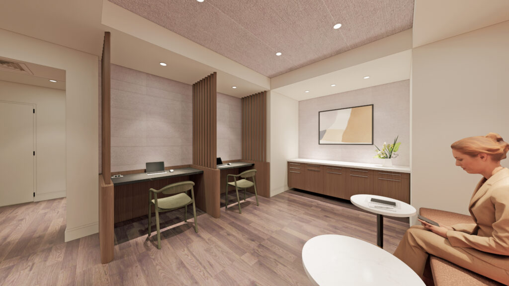 A modern office lounge with wooden floors, two workstations with chairs and laptops, a countertop with a framed abstract artwork above it, and a woman in a beige suit seated on a bench using her phone.