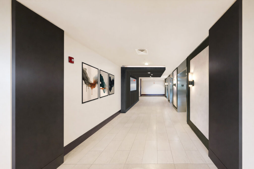 A modern hallway with light tiled floors, dark trim, three abstract paintings on the left wall, and wall sconces on the right. The hallway leads to elevators and doors at the far end.