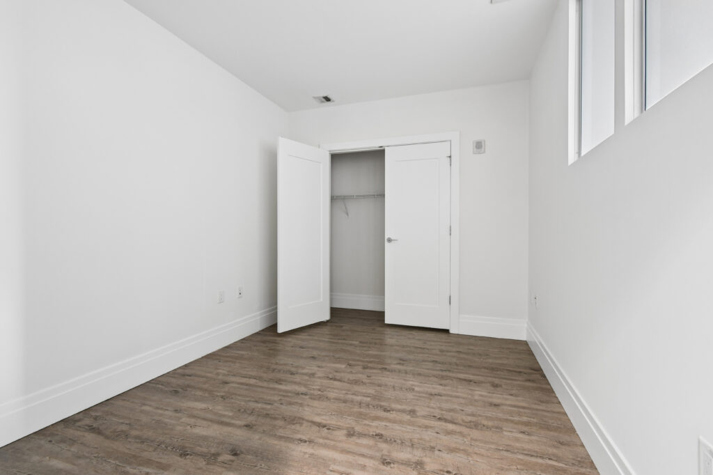 A bright, empty room with white walls, two white doors (one open to a closet), three high windows, and wood-style flooring. The space is minimal and modern.