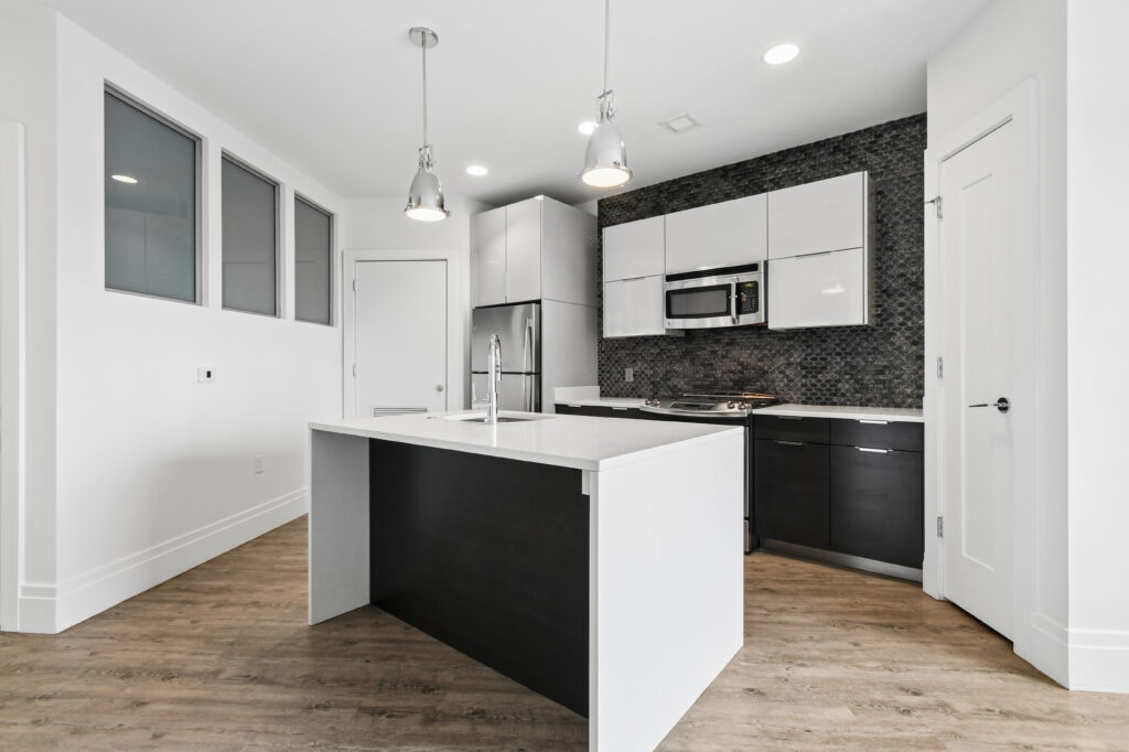 Modern kitchen with a white island, stainless steel appliances, black and white cabinets, black tile backsplash, pendant lights, and light wood flooring. Three frosted windows and two doors are visible.