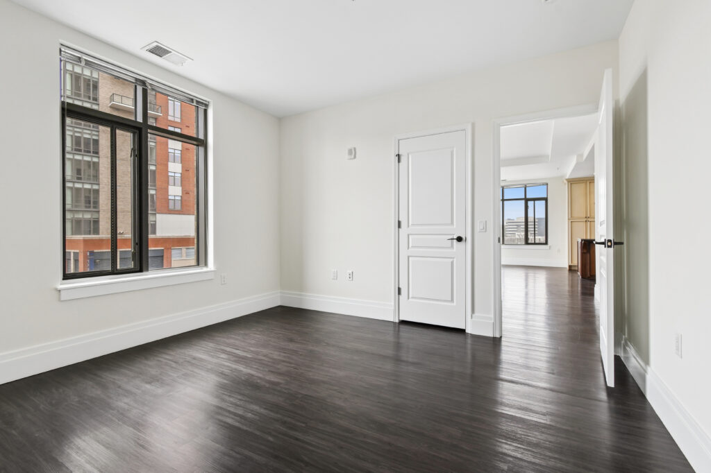 A bright, empty room with dark wood floors, white walls, a large window, and two doors; one door leads to another sunlit room with similar flooring and another large window.