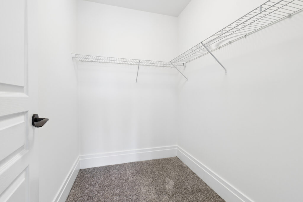 Empty walk-in closet with white walls, a white door, beige carpet, and two white wire shelves mounted on adjacent walls. The space is well-lit and clean, with no items stored inside.