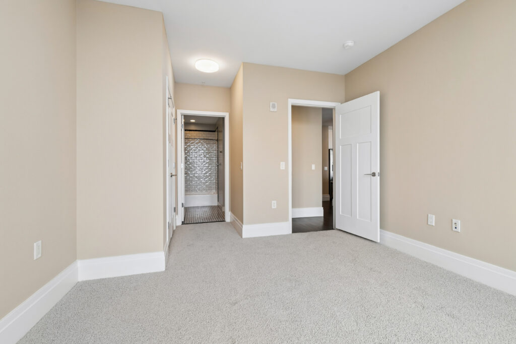A bright, empty bedroom with beige walls, white trim, light gray carpet, and an open door leading to a hallway. Another door reveals a bathroom with a tiled shower in the background.