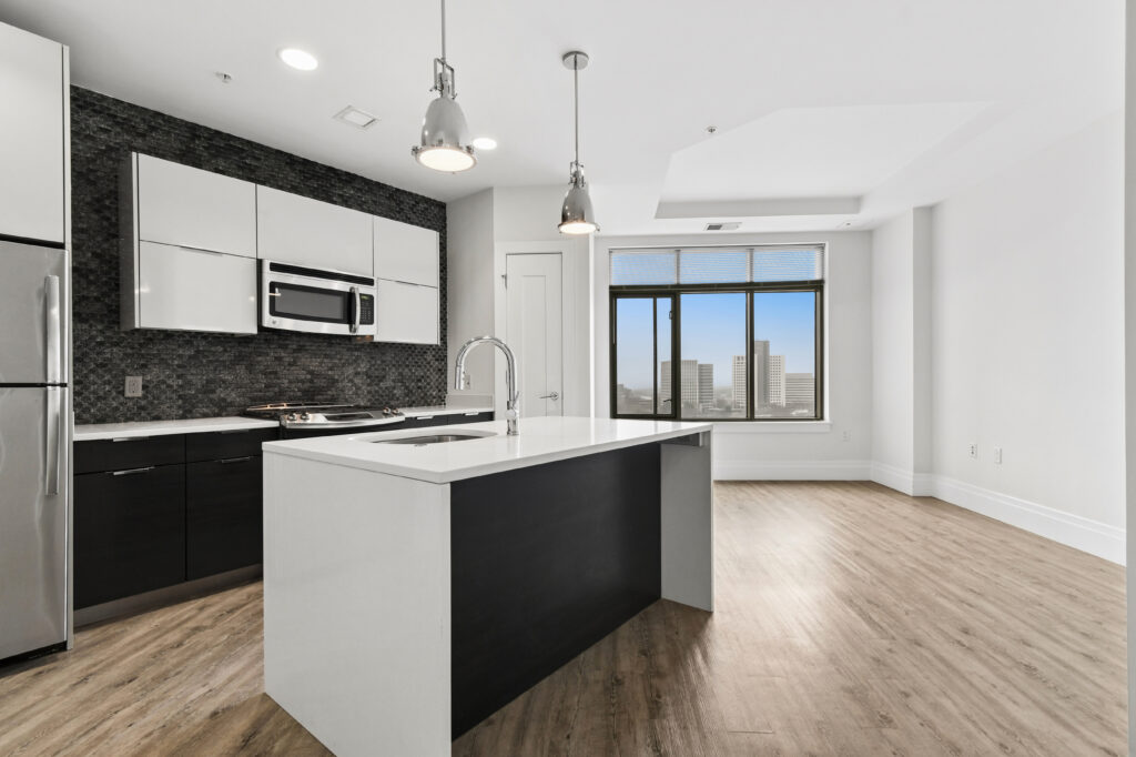 Modern kitchen with white countertops, black and white cabinets, stainless steel appliances, pendant lights, and wood flooring; large windows provide a city view and natural light.
