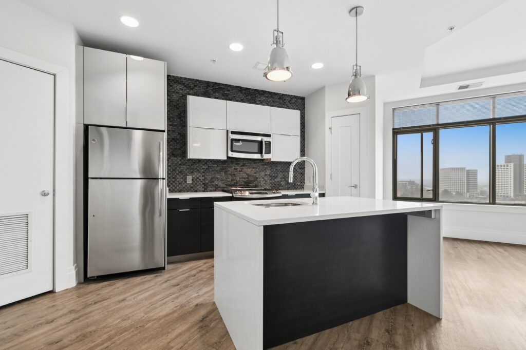 Modern kitchen with stainless steel appliances, white and black cabinets, a large island with a sink, wood flooring, pendant lights, and a large window with a cityscape view.
