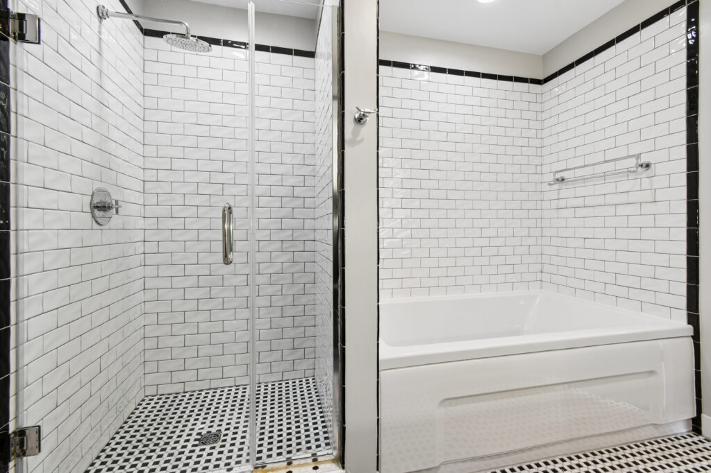 Modern bathroom with a walk-in glass shower next to a white bathtub. White subway tiles with black grout line the walls, and the floor features small black and white mosaic tiles.