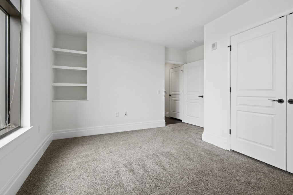 Empty, bright room with light-colored walls, carpeted floor, built-in shelves, a large window on the left, and two closed white doors on the right.