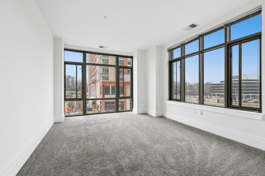 A bright, empty room with large floor-to-ceiling windows, gray carpet, and white walls, overlooking a street with construction and office buildings visible outside.