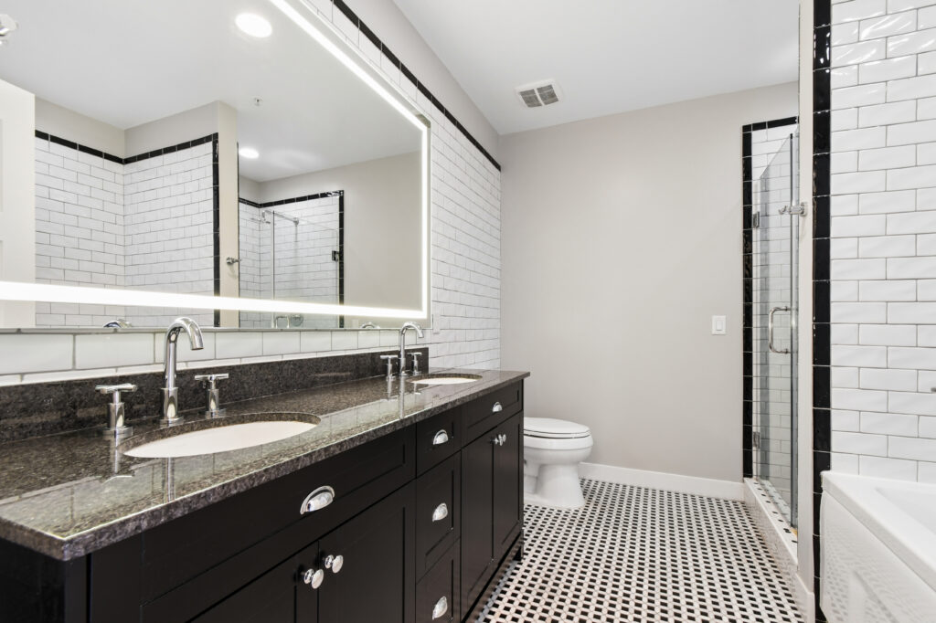 Modern bathroom with a black double vanity, white sink, large illuminated mirror, wall-mounted faucets, white subway tile walls, glass shower, toilet, and black-and-white patterned floor tiles.