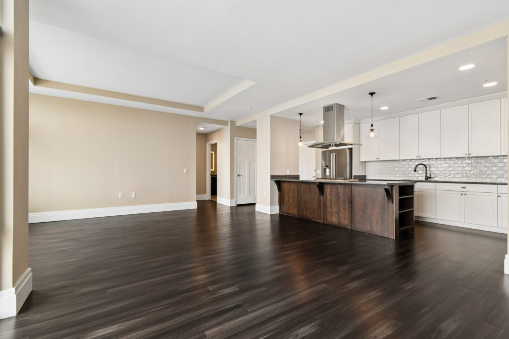 Modern, unfurnished open-concept living space with dark hardwood floors, beige walls, recessed lighting, and a kitchen featuring white cabinets, stainless steel appliances, and a dark wood island with pendant lights.