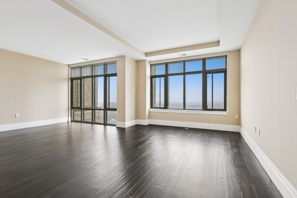 Spacious, empty room with dark wooden floors, beige walls, large windows, and a view of the city skyline with blue sky visible outside.